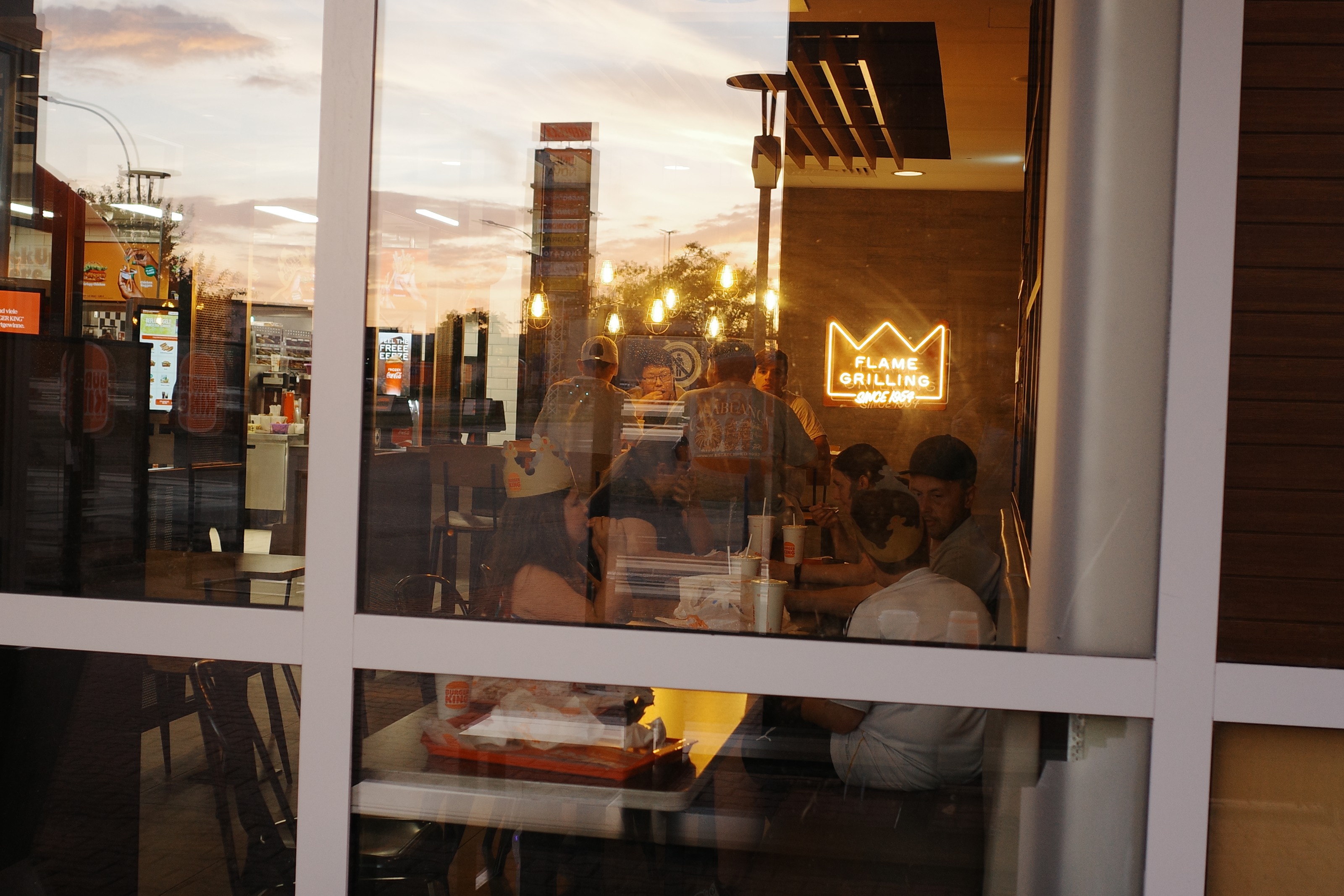 Diners behind a burger restaurant window at dusk, sunset reflected in the glass