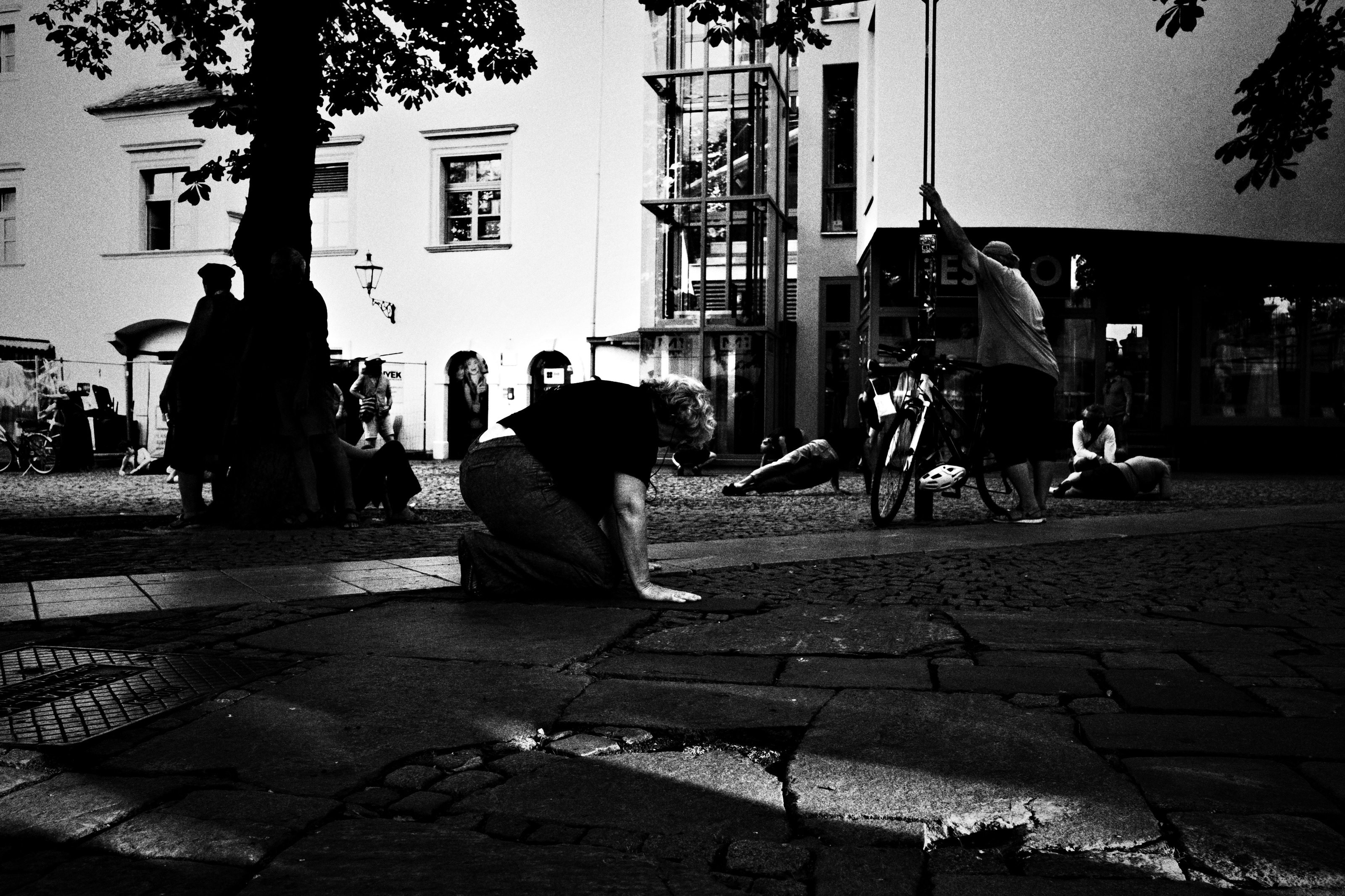 Black and white square at dusk, a woman crouched on the flagstones while others sit in the shade behind her