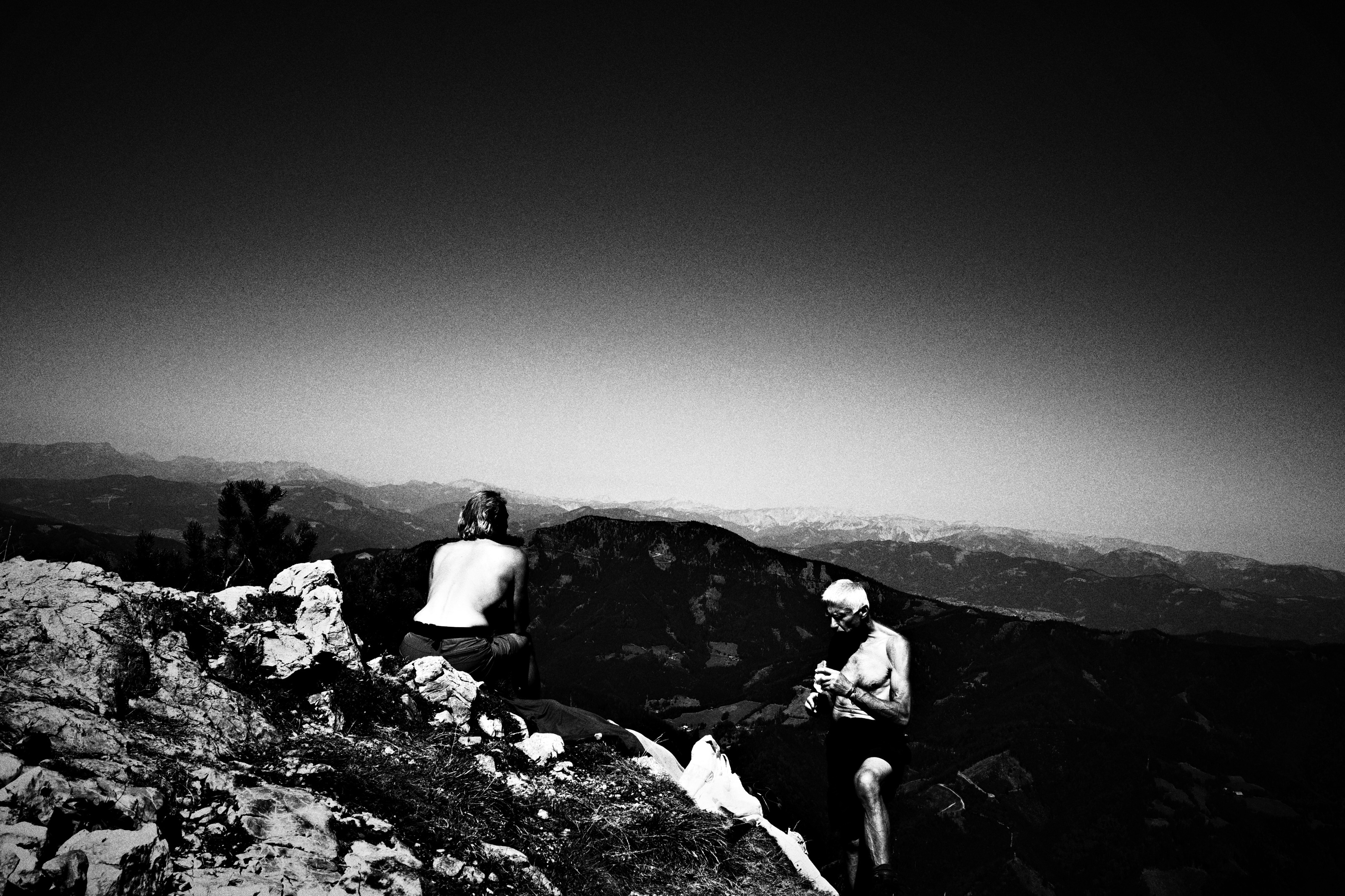 Two bare-chested hikers resting on a rocky summit above a long line of hazy peaks