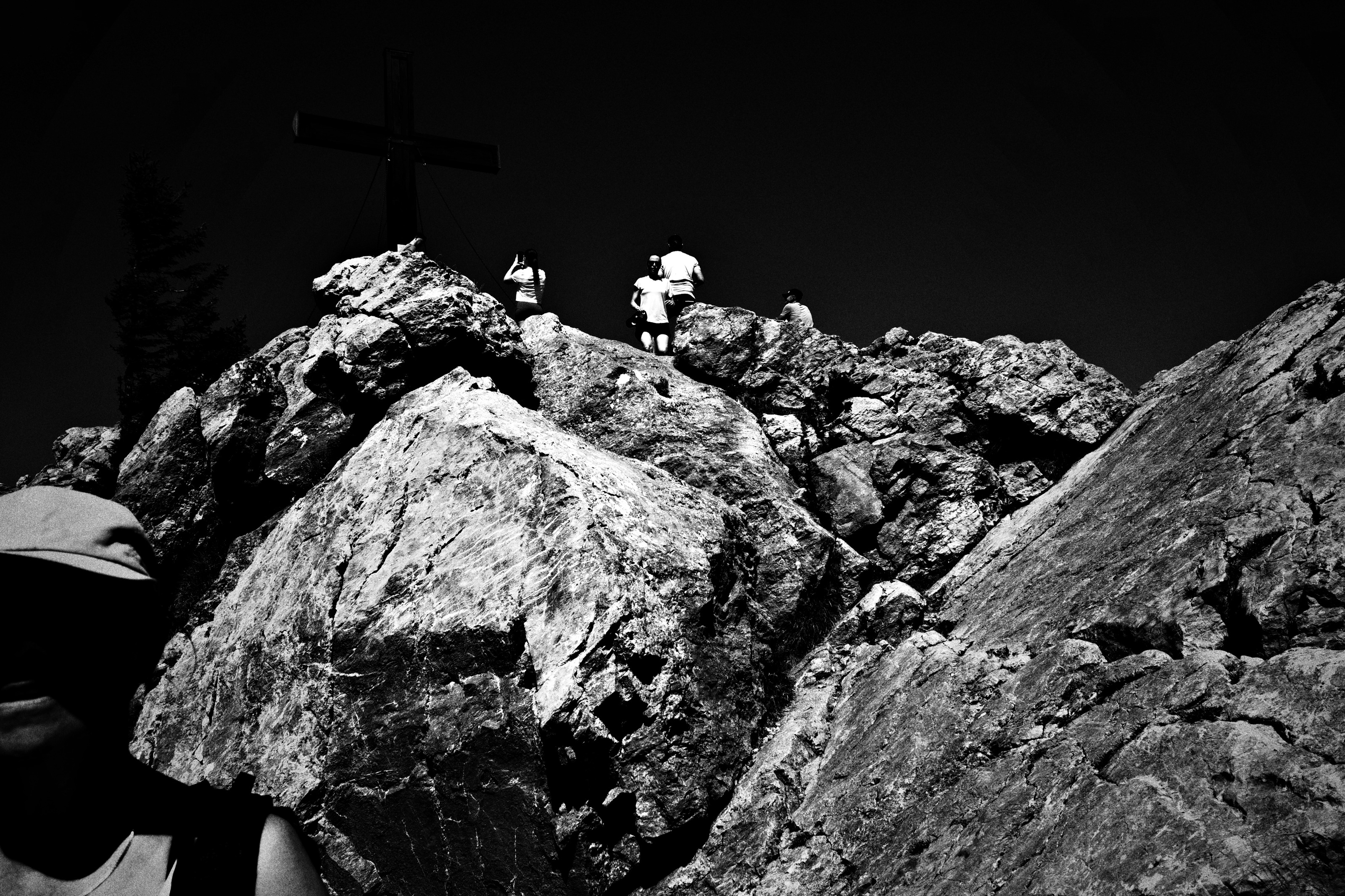 A summit cross on a pile of pale boulders, small figures climbing to it under a black sky
