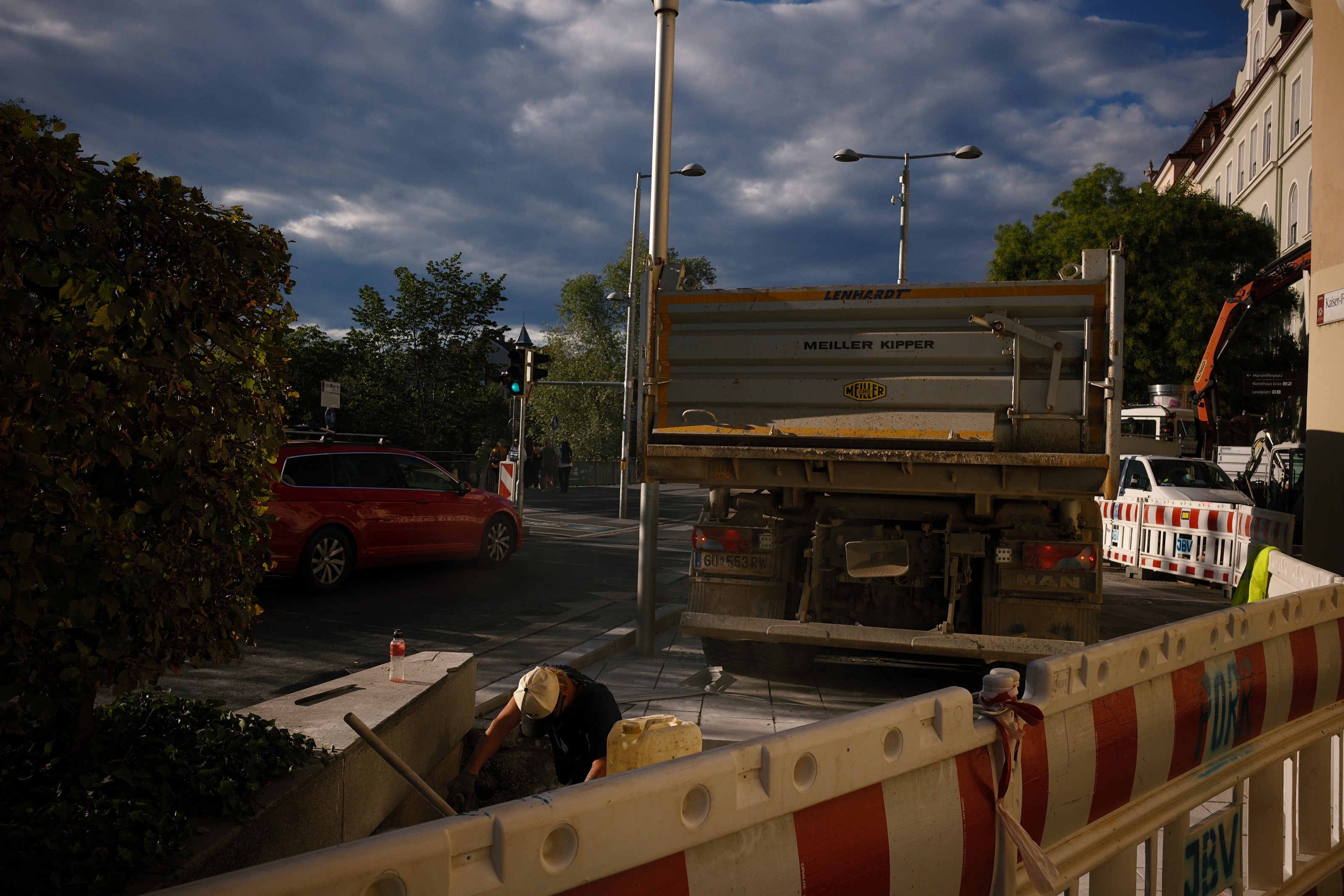 A tipper truck and red-white barriers at a crossing, a worker kneeling in an open trench under heavy cloud