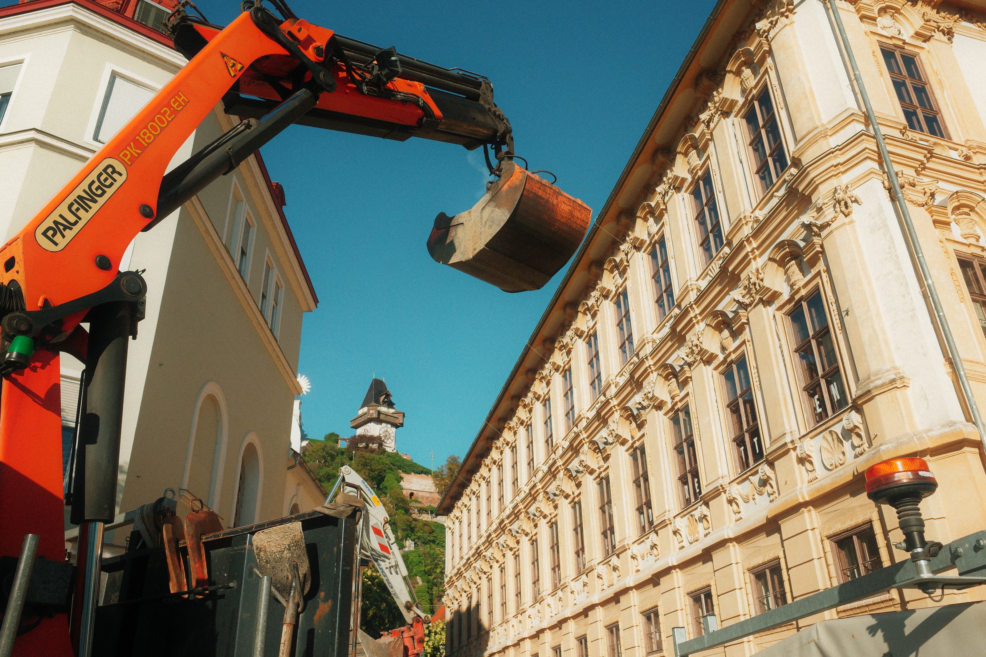An orange crane arm swinging a digger bucket over a street, the clock tower on its hill beyond