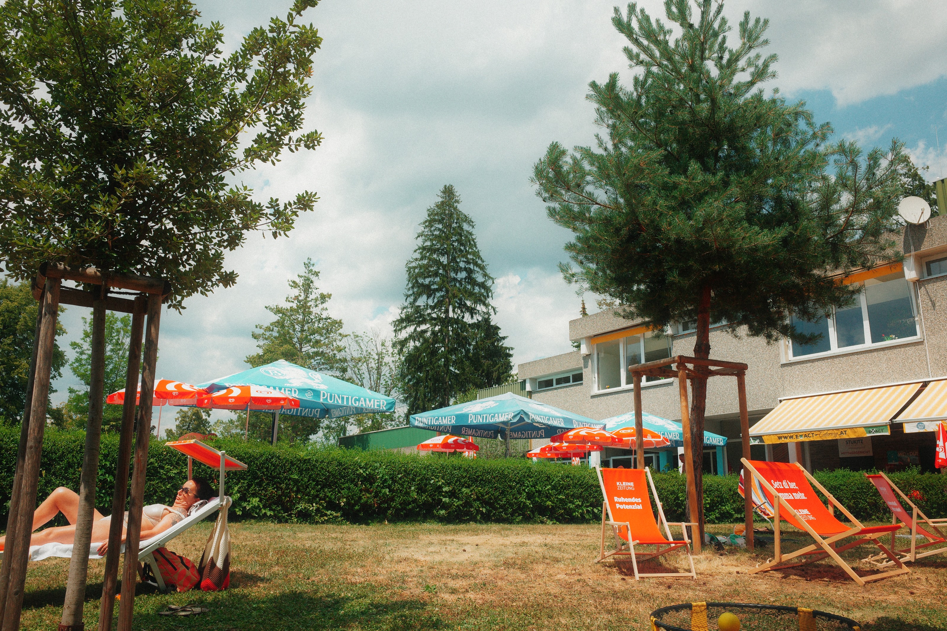 Empty striped deckchairs and beer umbrellas on a lawn, one sunbather in the shade
