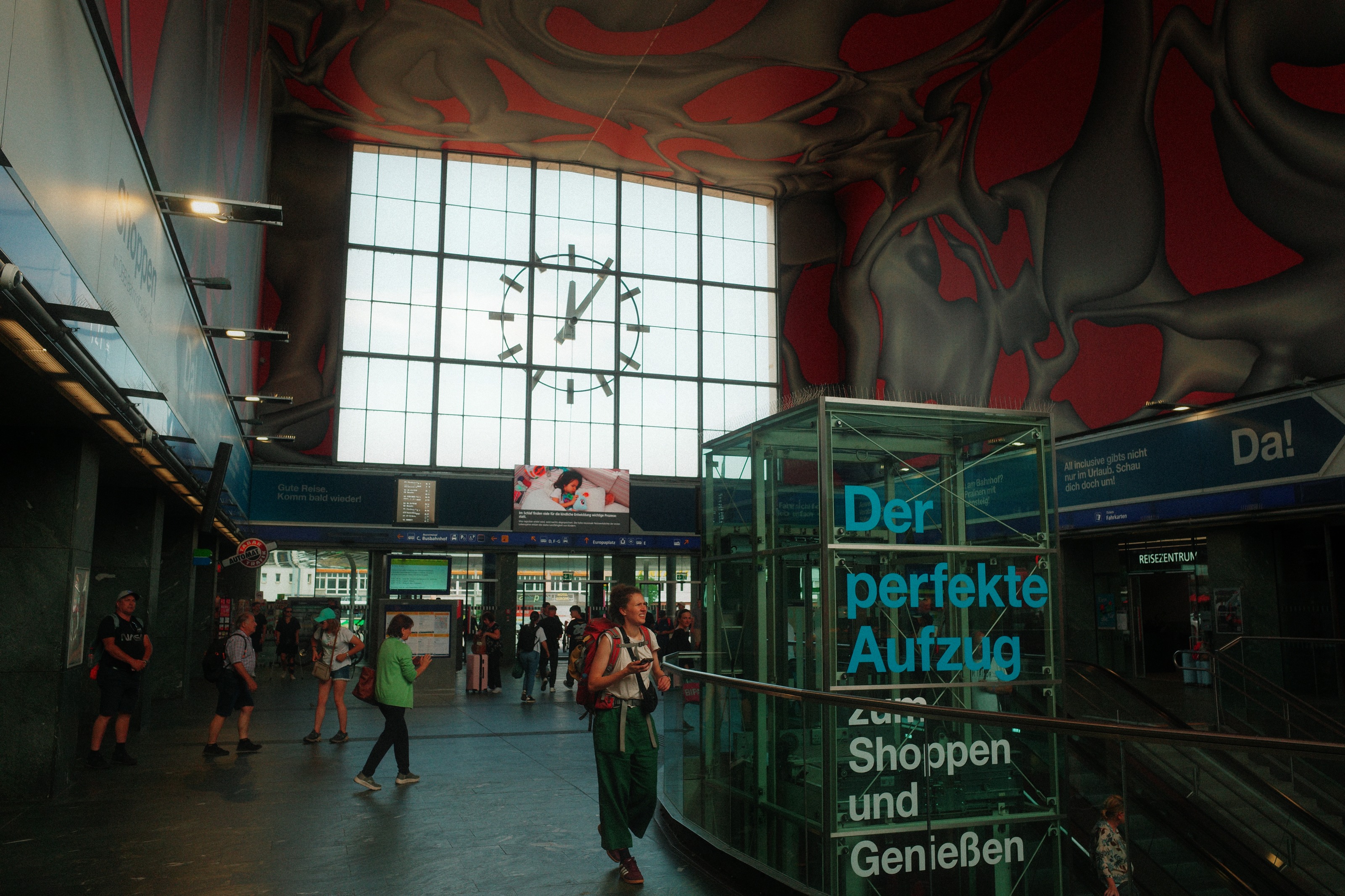 Station hall with a big clock in the window wall and a red mirrored ceiling sculpture above a glass lift