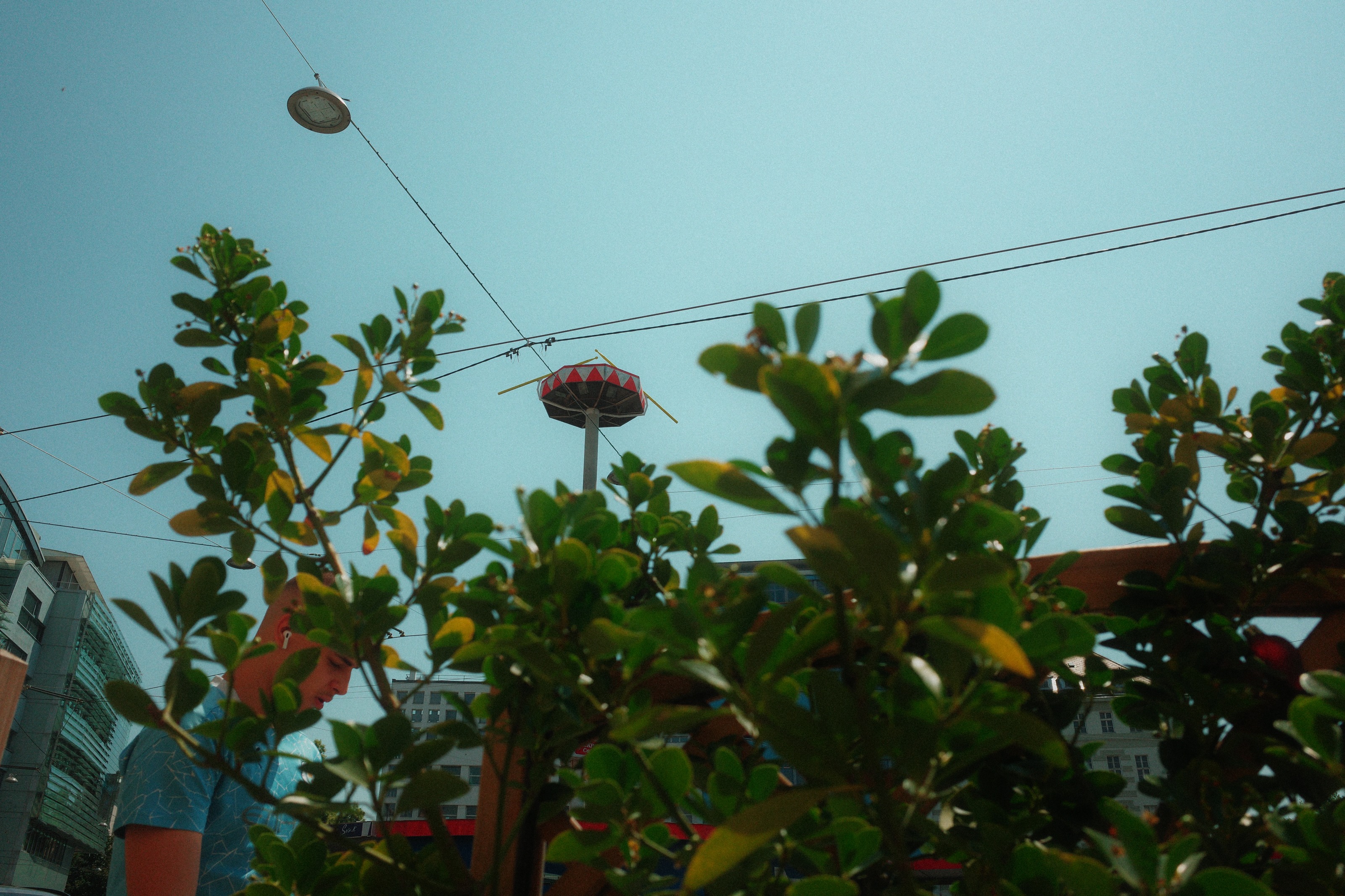 A fairground swing ride seen over hedge leaves and overhead wires against a pale blue sky